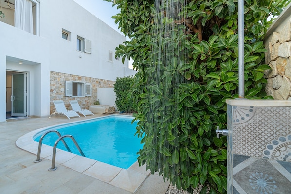 Una piscina con pared de piedra, un edificio blanco y un seto verde.