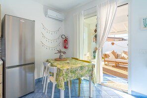 Una cocina con un refrigerador de acero inoxidable, una mesa de comedor con mantel floreado, sillas blancas y un extintor de incendios en la pared.