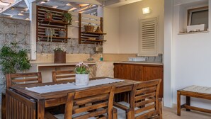 A wooden dining table with chairs, a kitchen area with shelves and potted plants, and a bench. - Apartment 'Falso Pepe Marzamemi' with Private Pool, Private Terrace and Air Conditioning (Marzamemi)