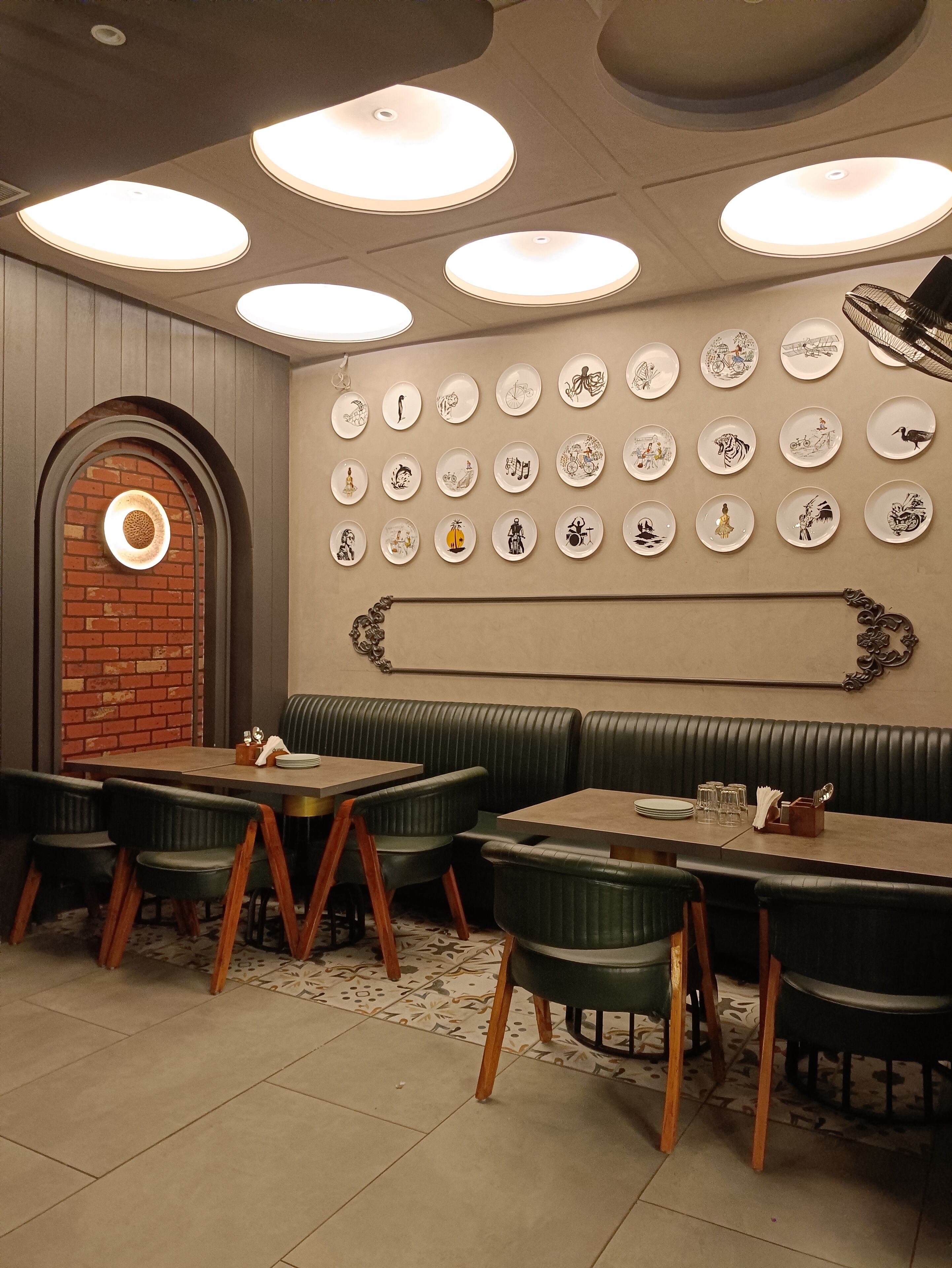 A restaurant interior with a brick wall, green upholstered booths, and a wall of decorative plates.