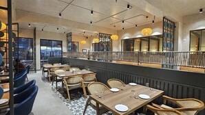 Een restaurant met houten tafels en rieten stoelen, een hoog plafond en hanglampen.