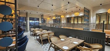 A restaurant with wooden tables and wicker chairs, a high ceiling, and pendant lighting.