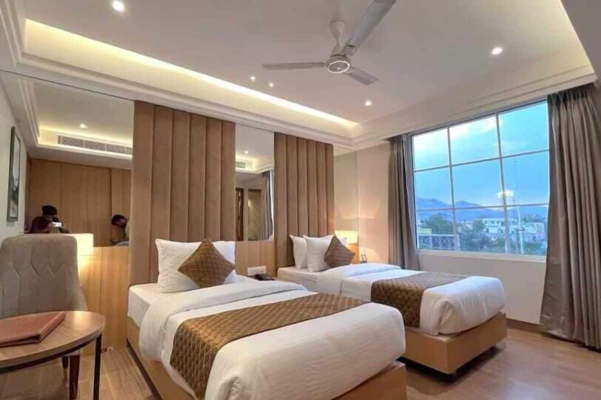 Une chambre d’hôtel avec deux lits, une petite table, une chaise, un ventilateur de plafond et une grande fenêtre donnant sur un beau paysage.