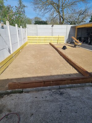 Een zanderig buitengebied met een houten bank, een geel hek en een betonnen muur.