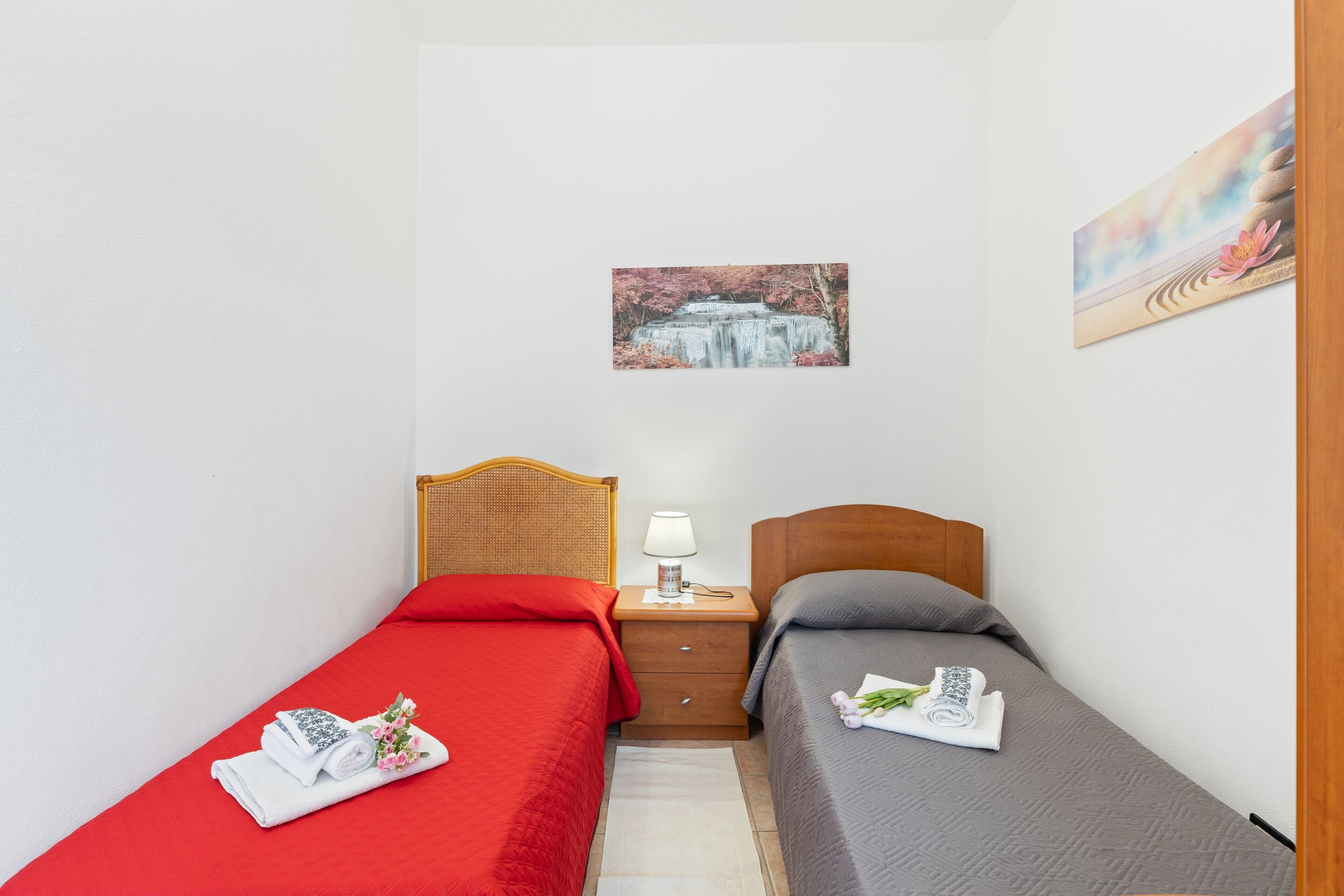 A room with two beds, a nightstand with a lamp, a small rug, and two wall-mounted pictures.