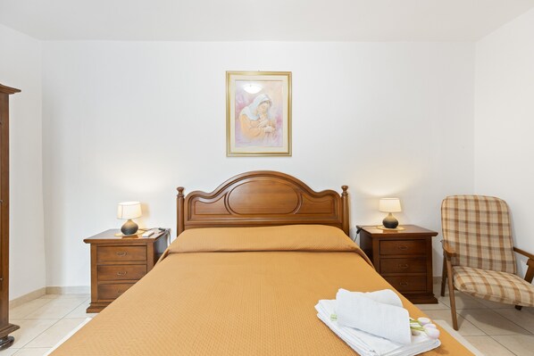 A neatly made bed with a headboard, two bedside tables with lamps, a chair, and a framed picture on the wall.