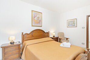 A hotel room with a wooden bed, a nightstand with a lamp, a chair, and two framed pictures on the wall.