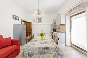 A dining area with a table, chairs, a red sofa, a refrigerator, a kitchen, and a chandelier.