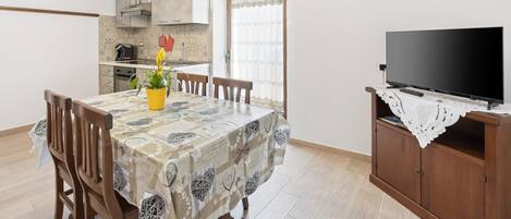 Un comedor con una mesa preparada para cuatro personas, un televisor sobre un soporte de madera y una cocina al fondo.
