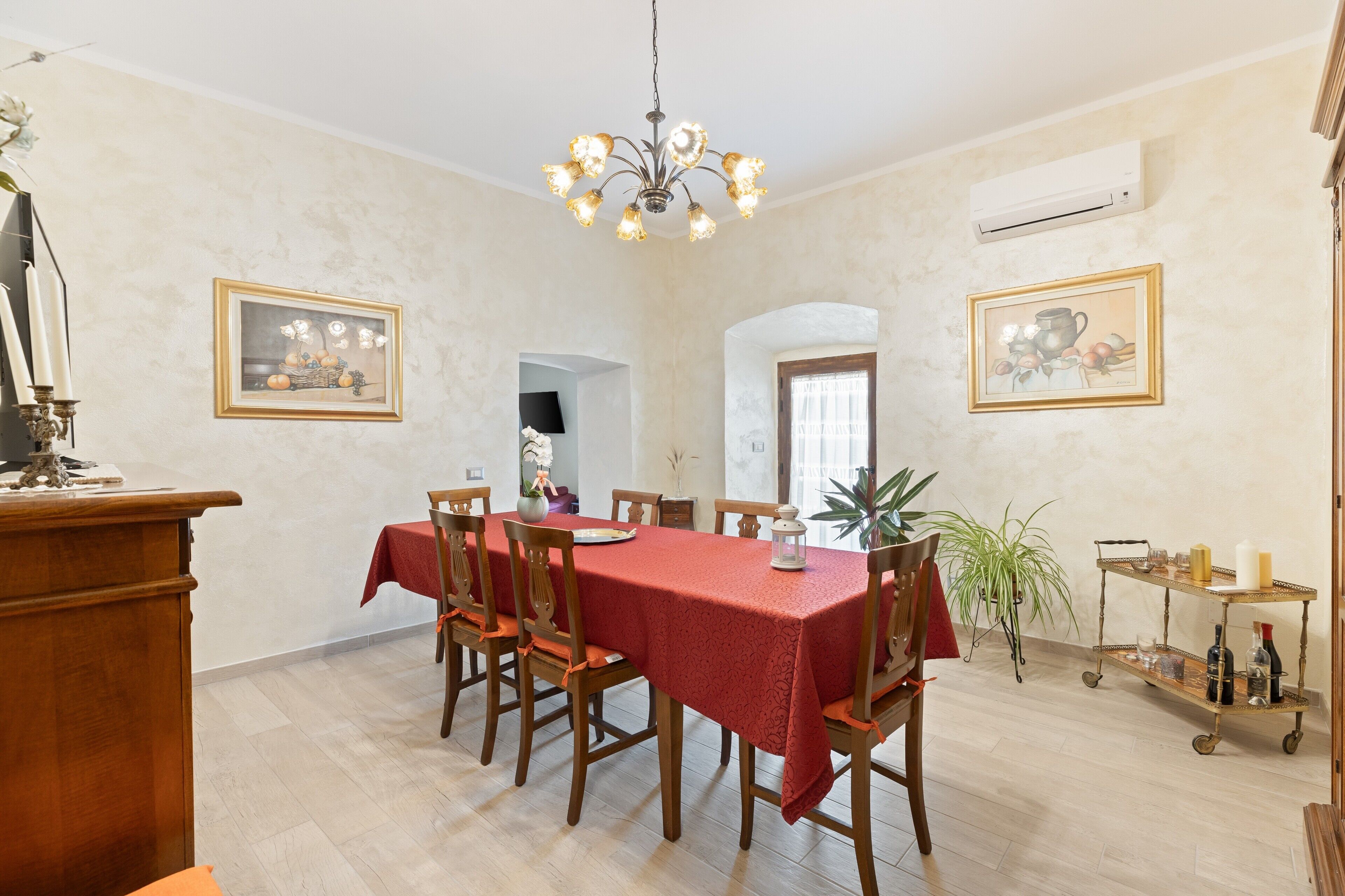 A dining room with a red tablecloth, wooden chairs, a chandelier, and wall-mounted air conditioning.