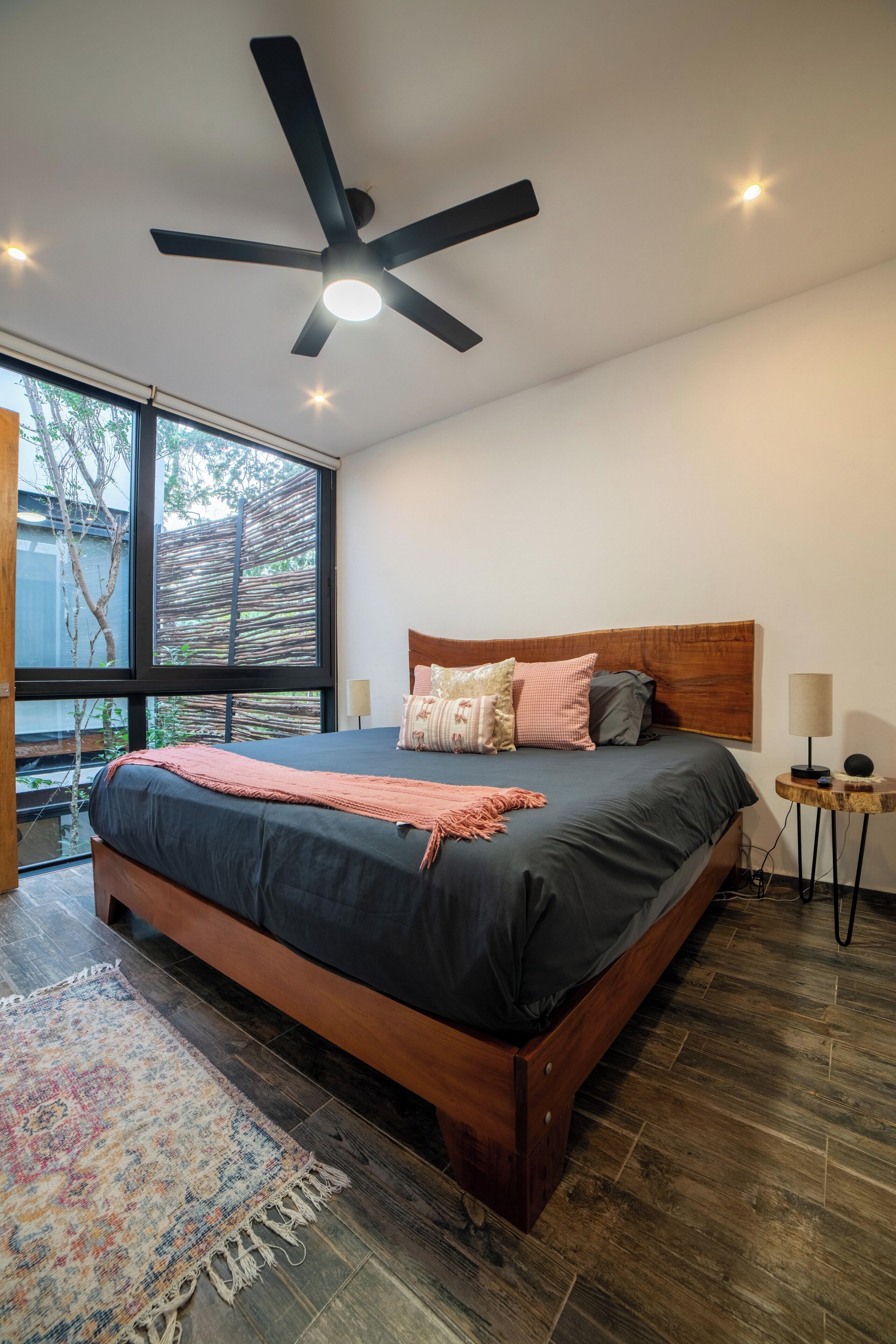 A modern bedroom with a large bed, a ceiling fan, a nightstand, and a view of trees through large windows.