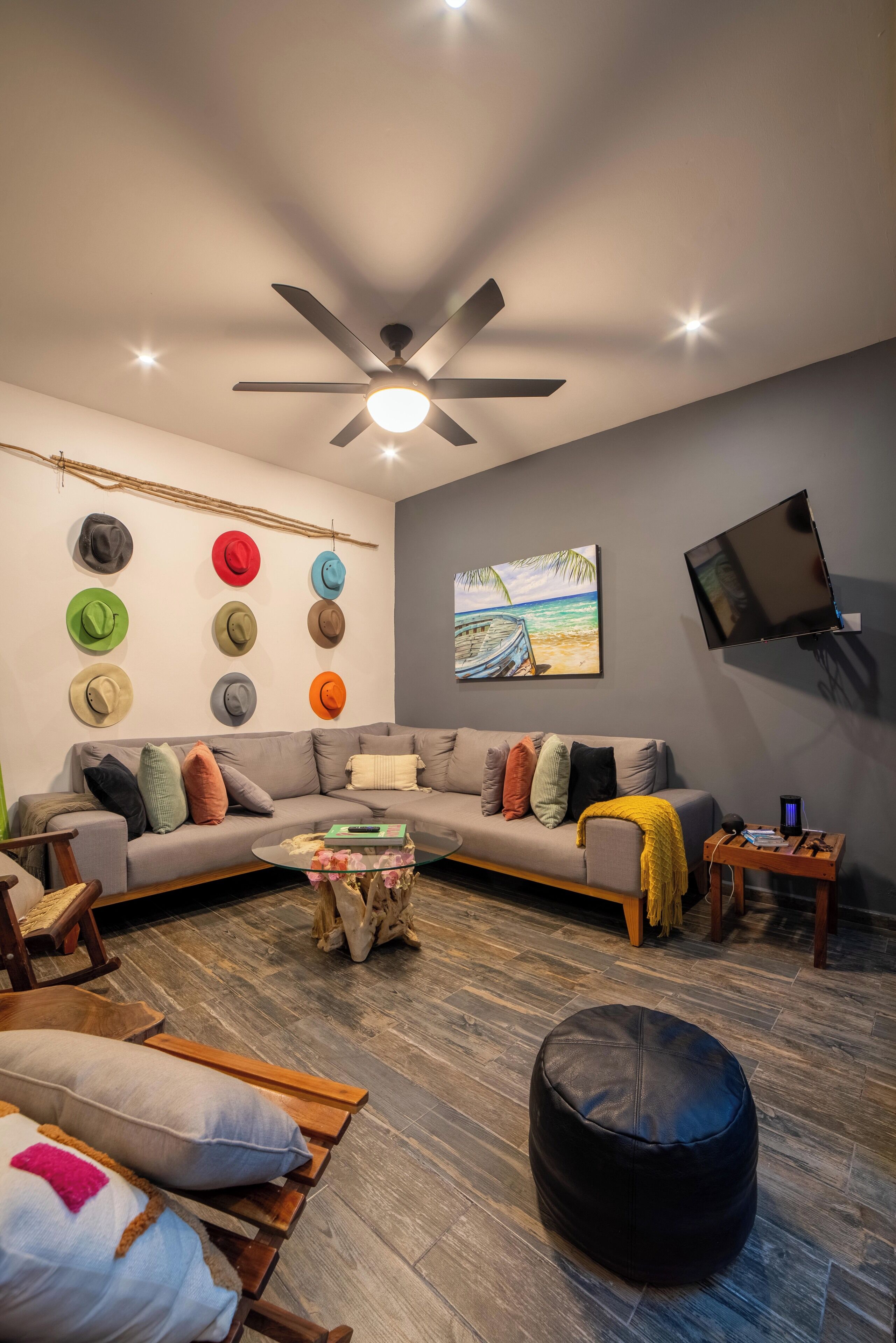 A modern living room with a sectional sofa, a coffee table, a black ottoman, a flat-screen TV mounted on the wall, and a ceiling fan.