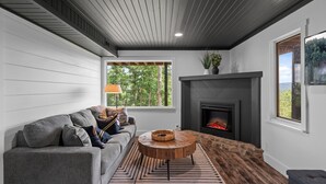 A modern living room with a grey sofa, a round wooden coffee table, a fireplace, and a view of trees through a window. - Panoramic Views! Wrap around Deck, Hot Tub & Grill (Morganton)