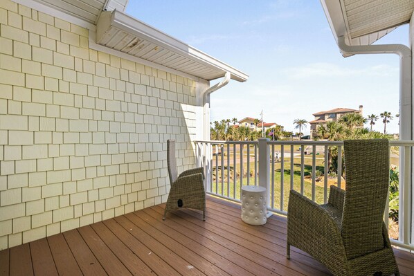 A covered porch with two wicker chairs and a small table, overlooking a landscaped area with palm trees and a distant building. - Flagler Beach BNB (Flagler Beach)