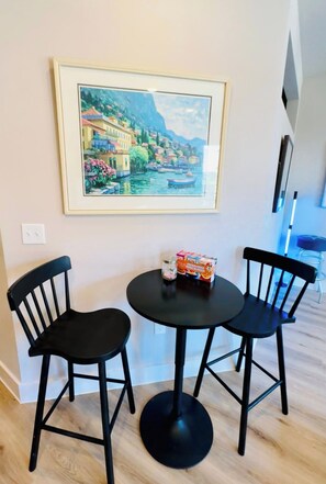A dining area with a round table and two chairs, a framed painting above, and a small table with a box and jar.