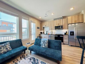 A modern apartment with a living area featuring two sofas, a kitchen with wooden cabinets, a stainless steel refrigerator, and a balcony with a view of buildings. - Peaceful North Austin Retreat Near The Domain (Austin)