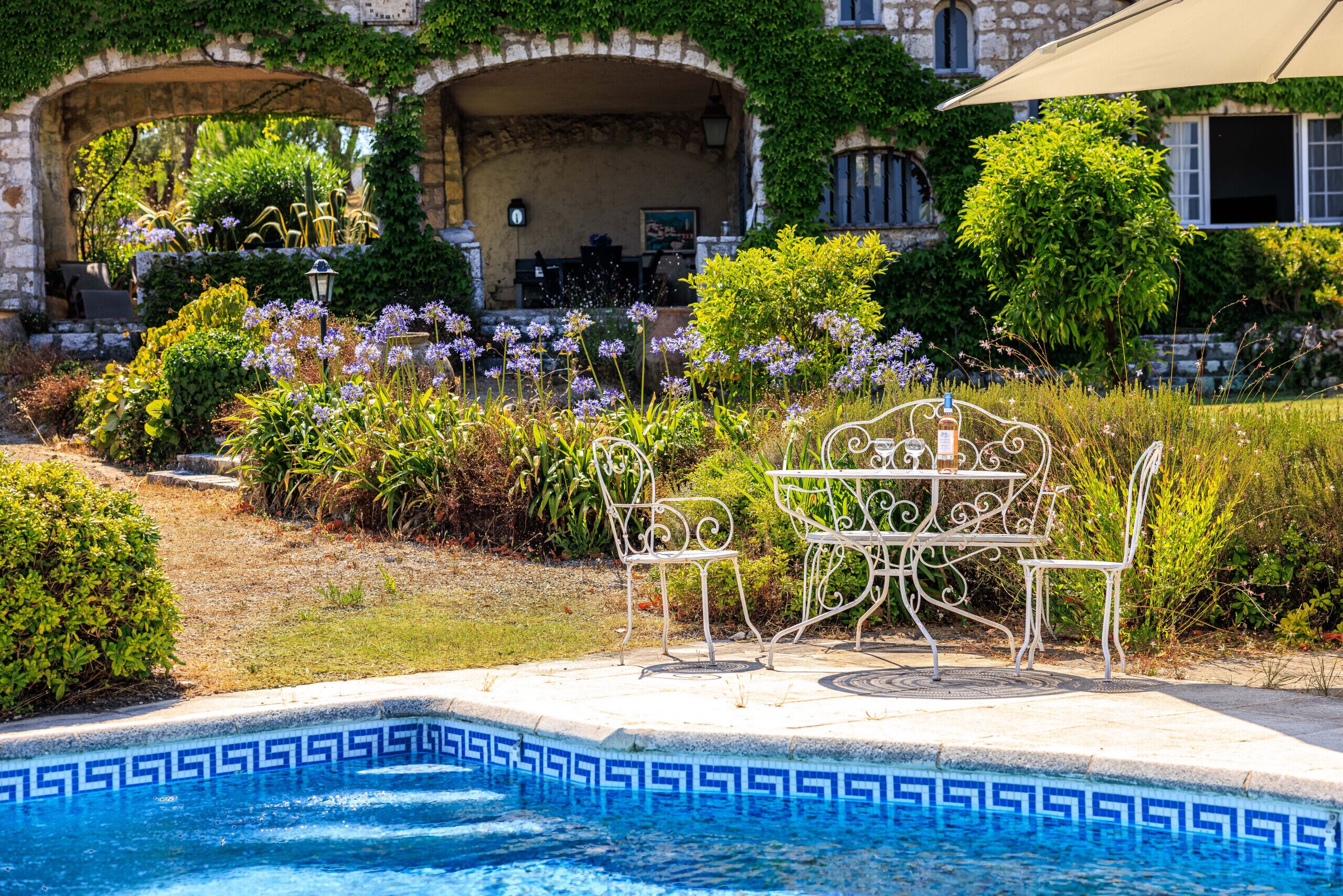 A pool area with a decorative edge, a white metal table and chairs, and a stone building with arches in the background.