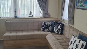 Un canapé beige agrémenté de coussins à motifs, une petite table avec une lampe et une fenêtre ornée de rideaux légers.