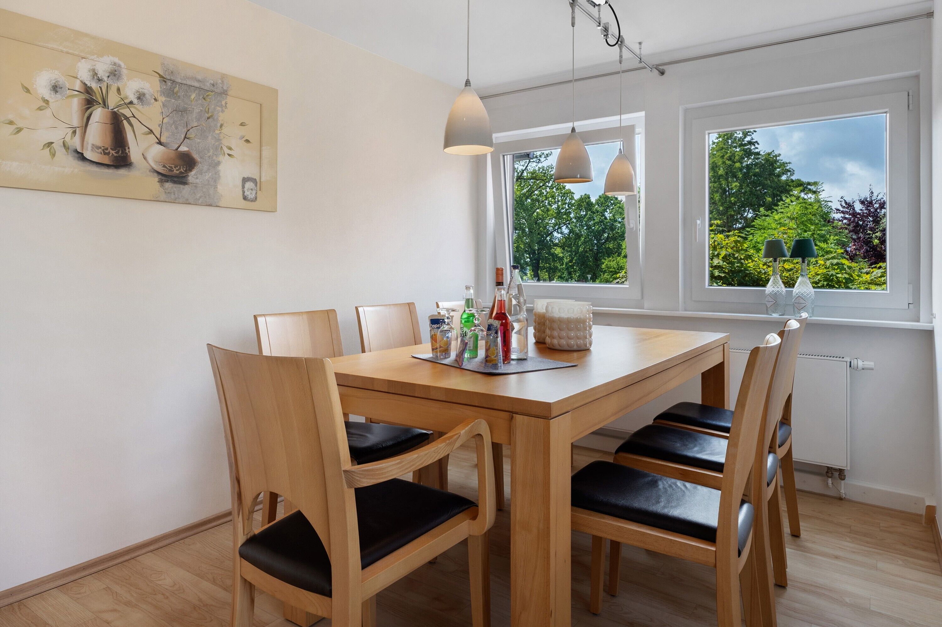 A dining area with a wooden table, chairs, pendant lights, and a window with a view of greenery.