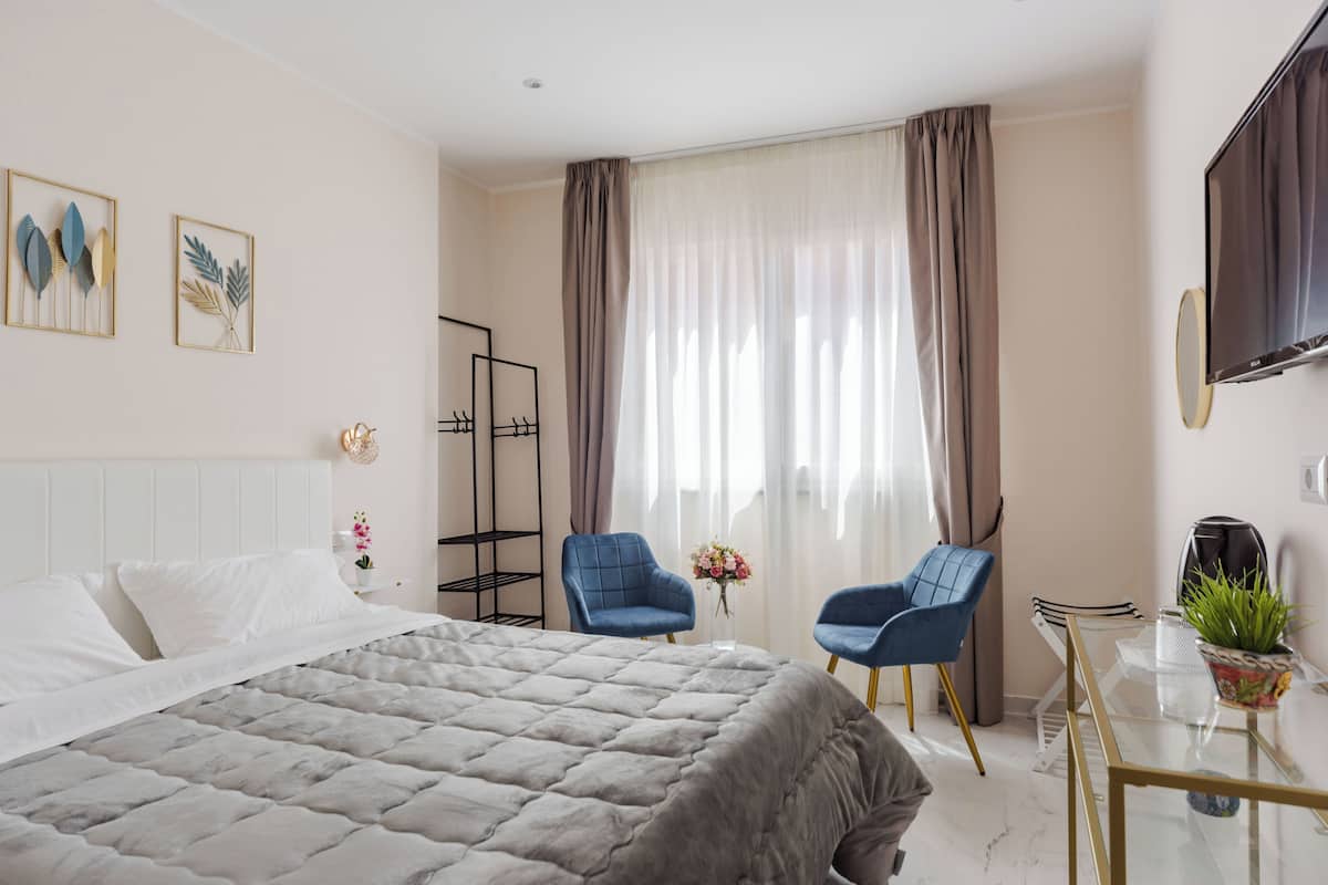 Ein modernes Hotelzimmer mit einem großen Bett, zwei blauen Stühlen, einem Glastisch, einem Fernseher und einem Fenster mit Vorhängen.