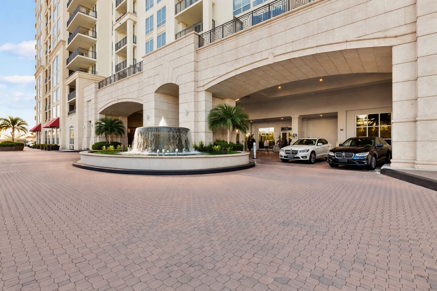 A hotel entrance with a fountain, palm trees, and cars parked outside.
