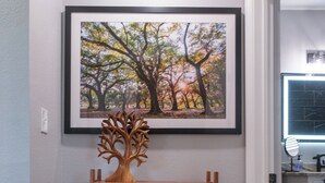 Una consola de madera con un árbol decorativo, un cuenco y un florero con flores frente a una pared con un cuadro enmarcado encima.