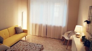 Ein Wohnzimmer mit gelbem Sofa, einem gemusterten Hocker, einem weißen Sessel, einer Lampe und einem großen Fenster mit transparenten Vorhängen.
