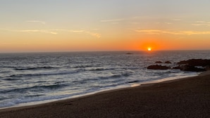 A beach with the sun setting over the ocean, waves gently lapping the shore, and rocky outcrops in the distance. - Charming Duplex in Foz do Douro | Near Passeio Alegre & the Sea (Porto)