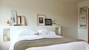 A neatly made bed with white linens and a gray throw, a bedside table with books and a vase of flowers, and a wall with framed pictures and a small shelf. - Charming Duplex in Foz do Douro | Near Passeio Alegre & the Sea (Porto)