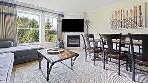 A living room with a sofa, a coffee table, a fireplace, a TV, and a dining area with chairs and a table. - Charming 2-Bedroom Ski-In/Ski-Out at Jackson Gore (Ludlow)