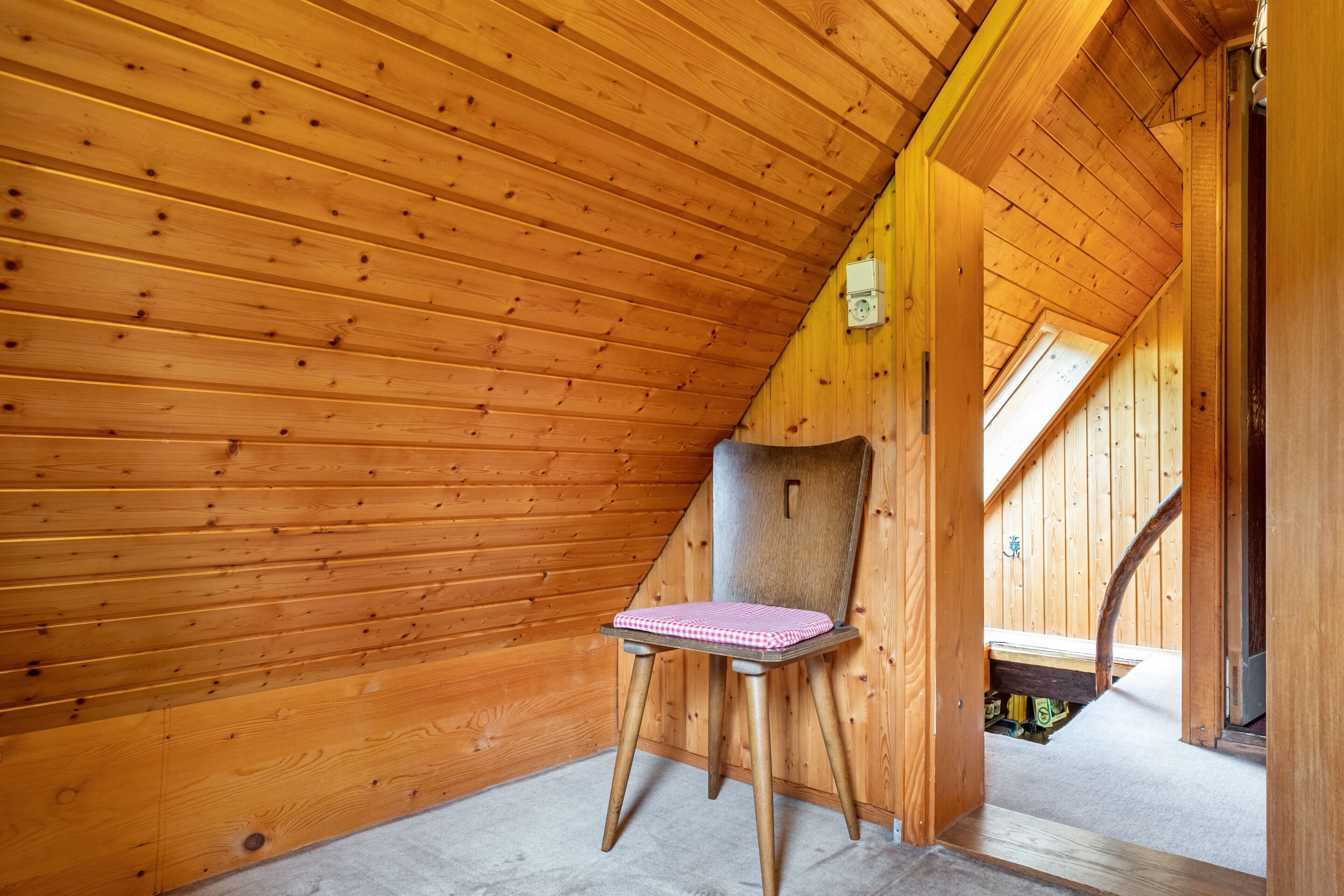 Un intérieur en bois avec un plafond en pente, une chaise dotée d’un coussin à carreaux et un escalier apparent menant à un niveau supérieur.