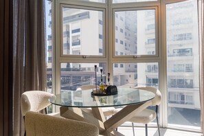 Et spisebord med glasplade, fire stole, en bakke med flasker og udsigt til høje bygninger gennem store vinduer.