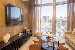 A modern living room with a large TV, a sofa, a coffee table, and a view of the city through large windows.