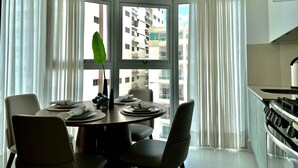 Un coin repas avec une table ronde dressée pour quatre personnes, une plante en pot et une vue sur le paysage urbain à travers de grandes fenêtres.