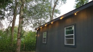 A dark wooden cabin with two windows and string lights along the roof edge.