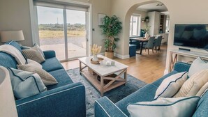 A spacious living room with a large sofa set, a coffee table, and a television. There is a sliding glass door leading to an outdoor area.