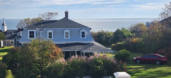 A two-story house with a gabled roof and a covered porch, surrounded by lush greenery and a view of the sea.