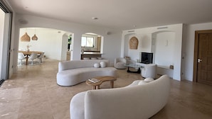A spacious living area with a large L-shaped sectional sofa, a small coffee table, and a television. There is a dining area with a wooden table and chairs visible through an archway. - New 5*star villa - Gulf of Saint-Tropez  - Beautiful sea view - (Cavalaire-sur-Mer)