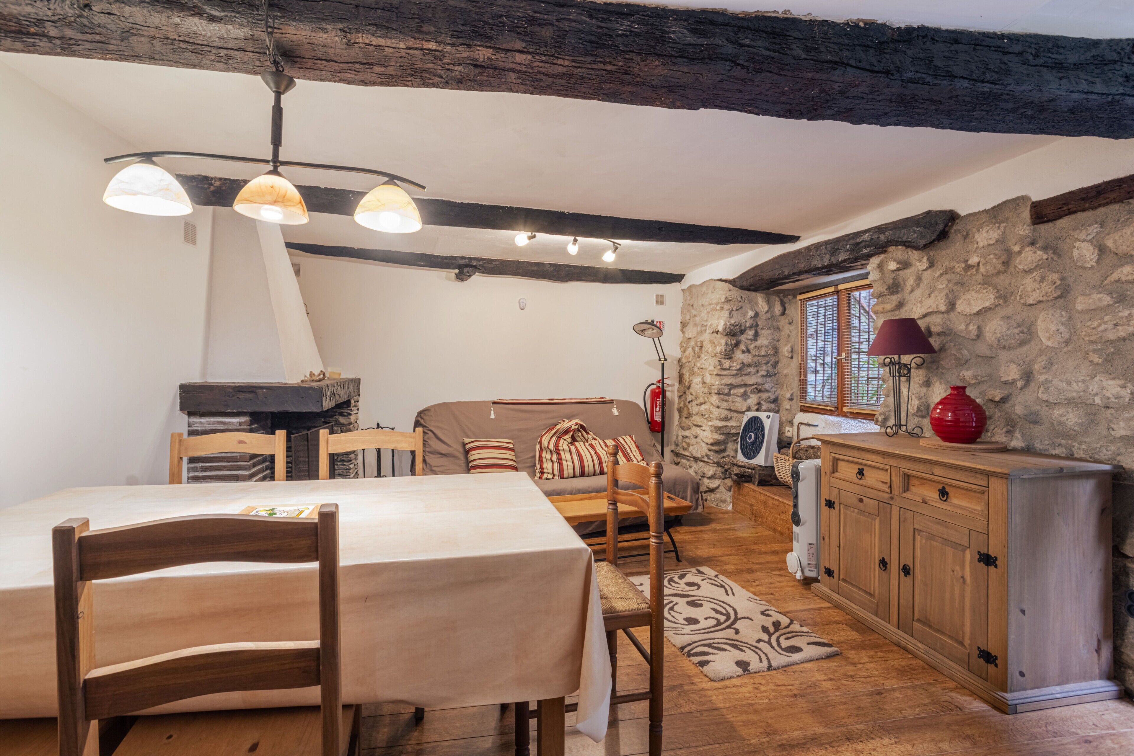 Un interior rústico con vigas a la vista, una pared de piedra, una zona de comedor con mesa y sillas, un sofá y un aparador de madera.