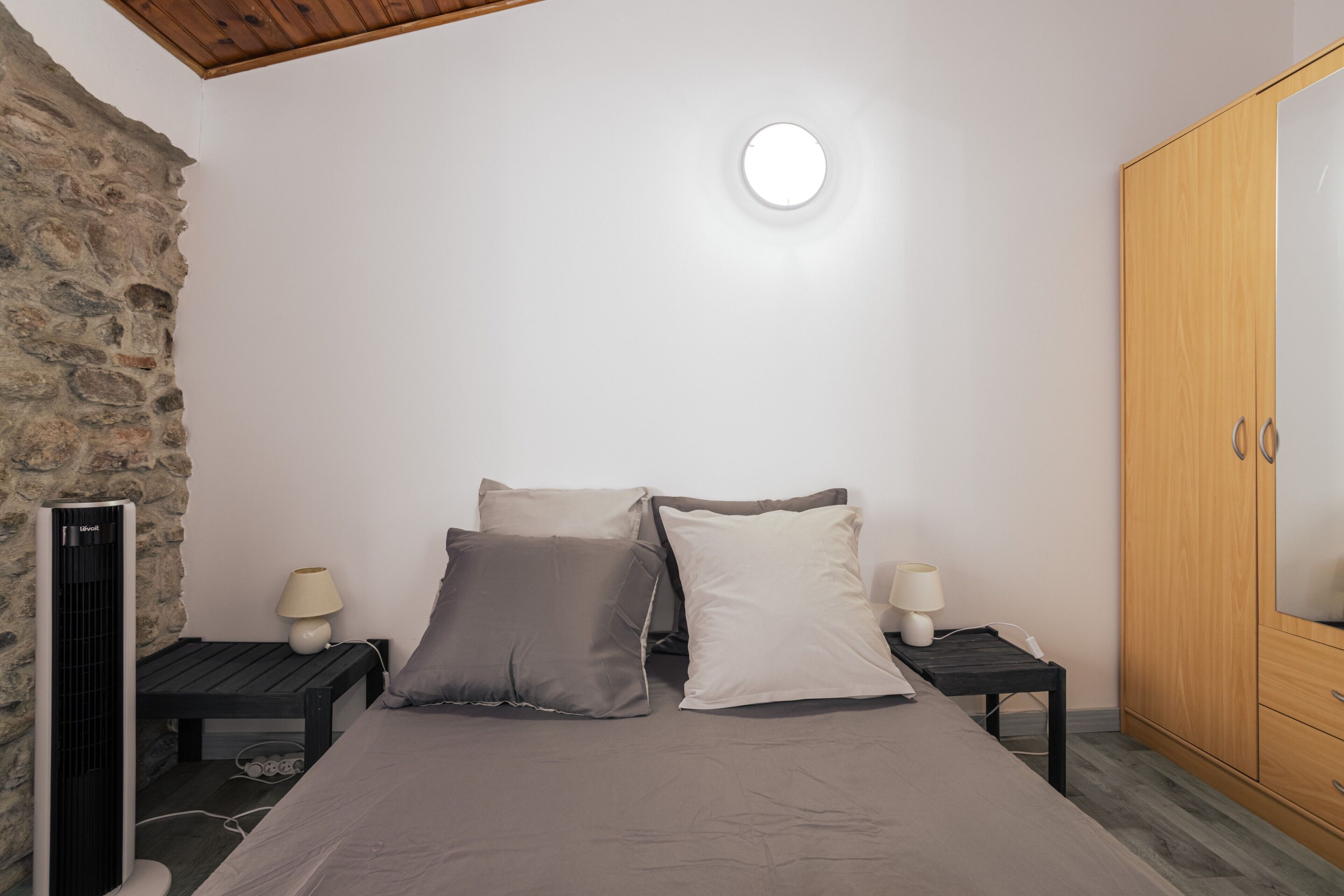 A bedroom with a bed, two bedside tables with lamps, a wardrobe, a stone wall, and a ceiling light.