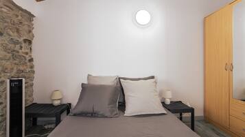 A bedroom with a bed, two bedside tables with lamps, a wardrobe, a stone wall, and a ceiling light.