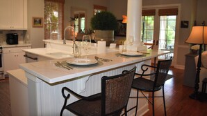 A kitchen with a central island, white cabinetry, and a dining area with two chairs. - Live Oaks Cottage - Golf Course Views & Lowcountry Charm in Bluffton, SC (Bluffton)