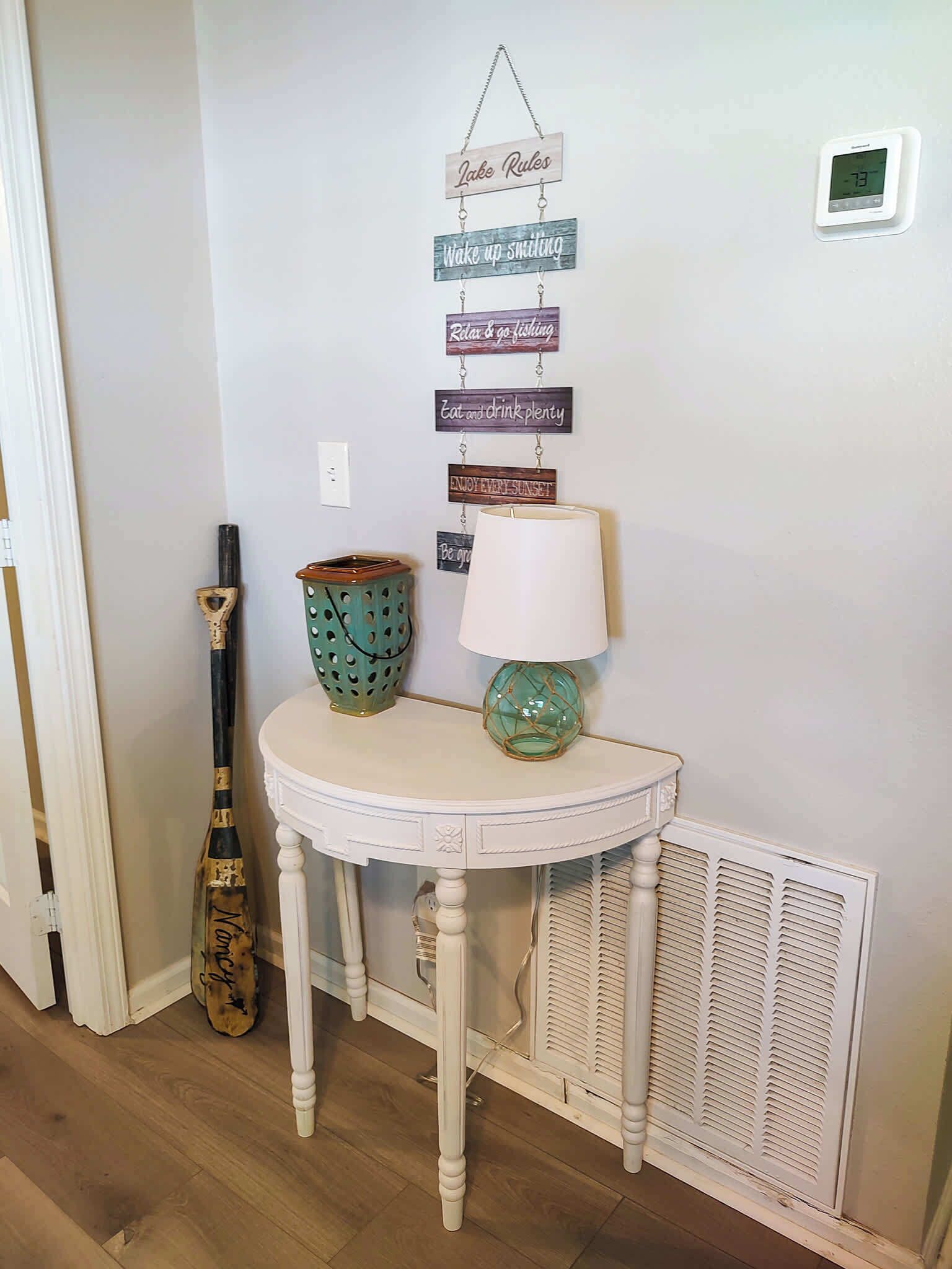 A white table with a lamp and decorative items, a wall-mounted sign with motivational phrases, and a vintage guitar leaning against the wall.