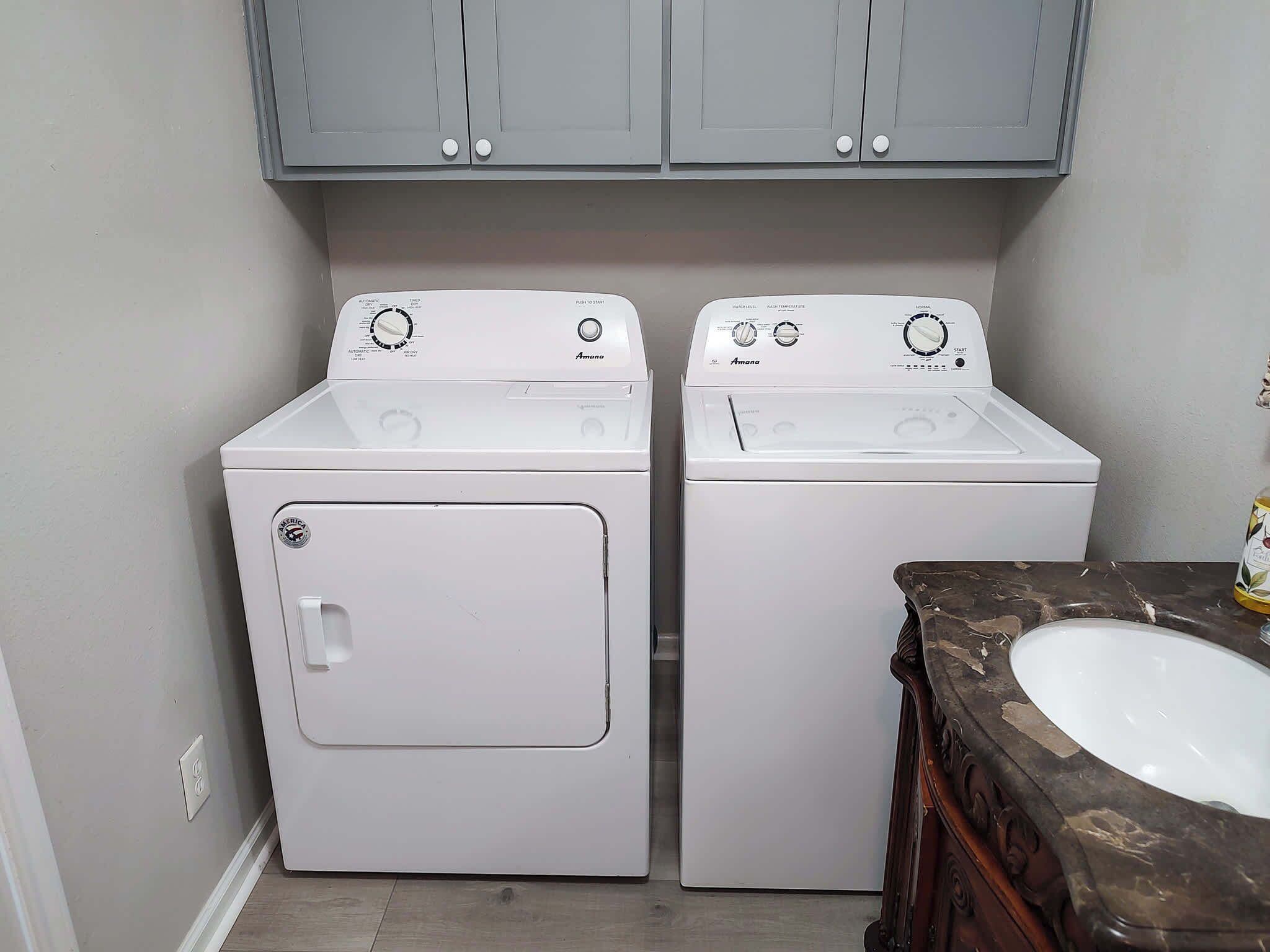 A laundry room with two white front-loading washers and dryers, grey cabinets above, and a marble sink with a wooden cabinet.