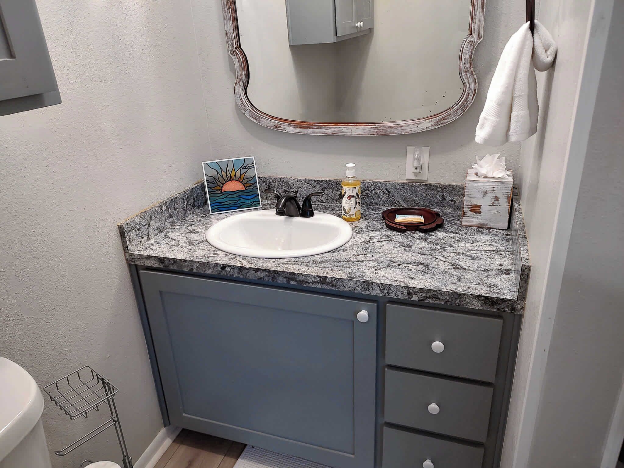 A bathroom with a white sink, a mirror with a decorative frame, a towel hanging on the wall, and a soap dispenser on the countertop.