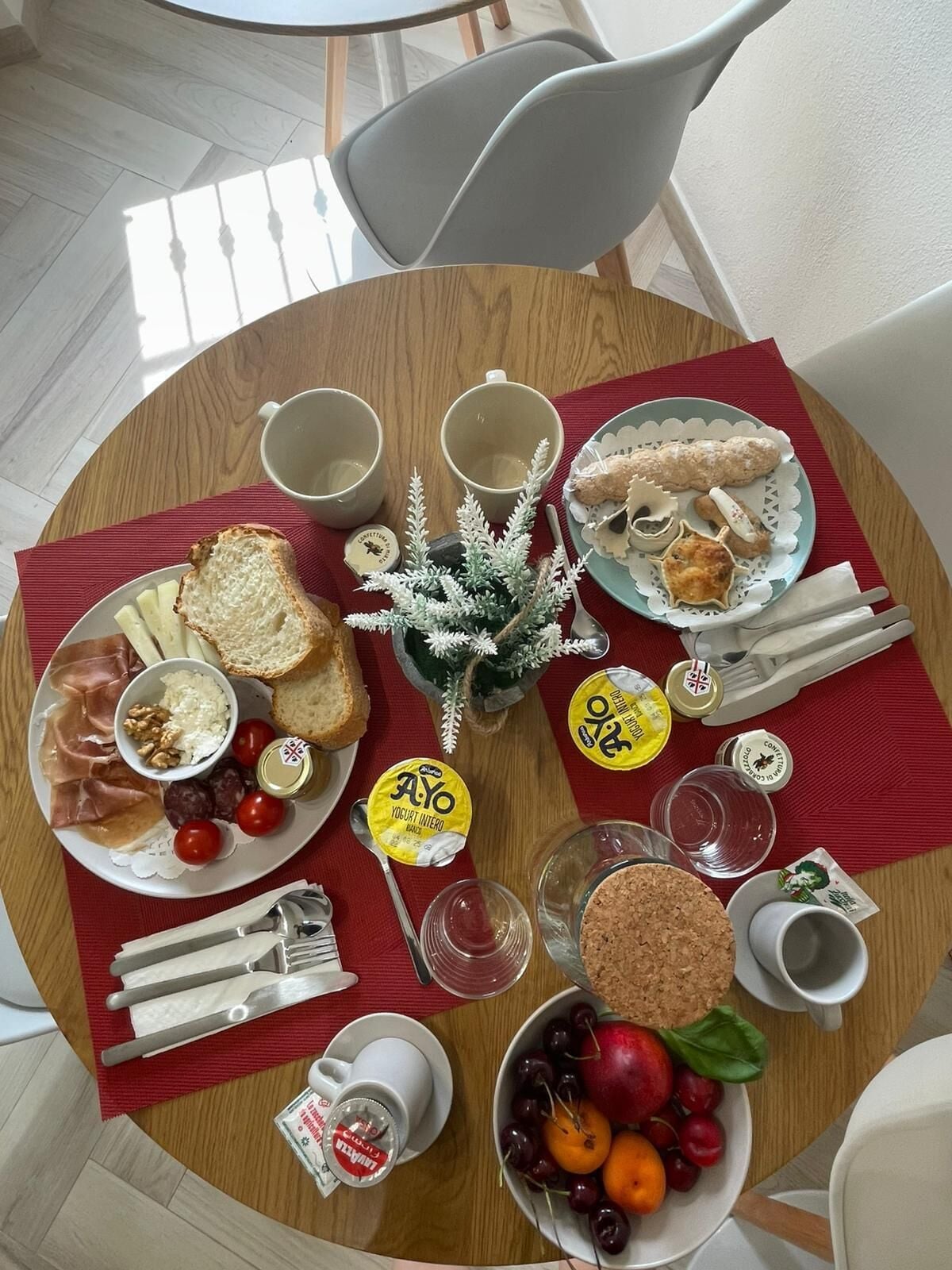A round wooden table with a red placemat, a small potted plant, and various food items including bread, cheese, and fruits.