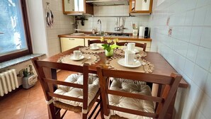 Un comedor con una mesa de madera dispuesta para cuatro personas, rodeada de sillas con asientos acolchados. Se observa una cocina en el fondo, con azulejos blancos y armarios de madera.