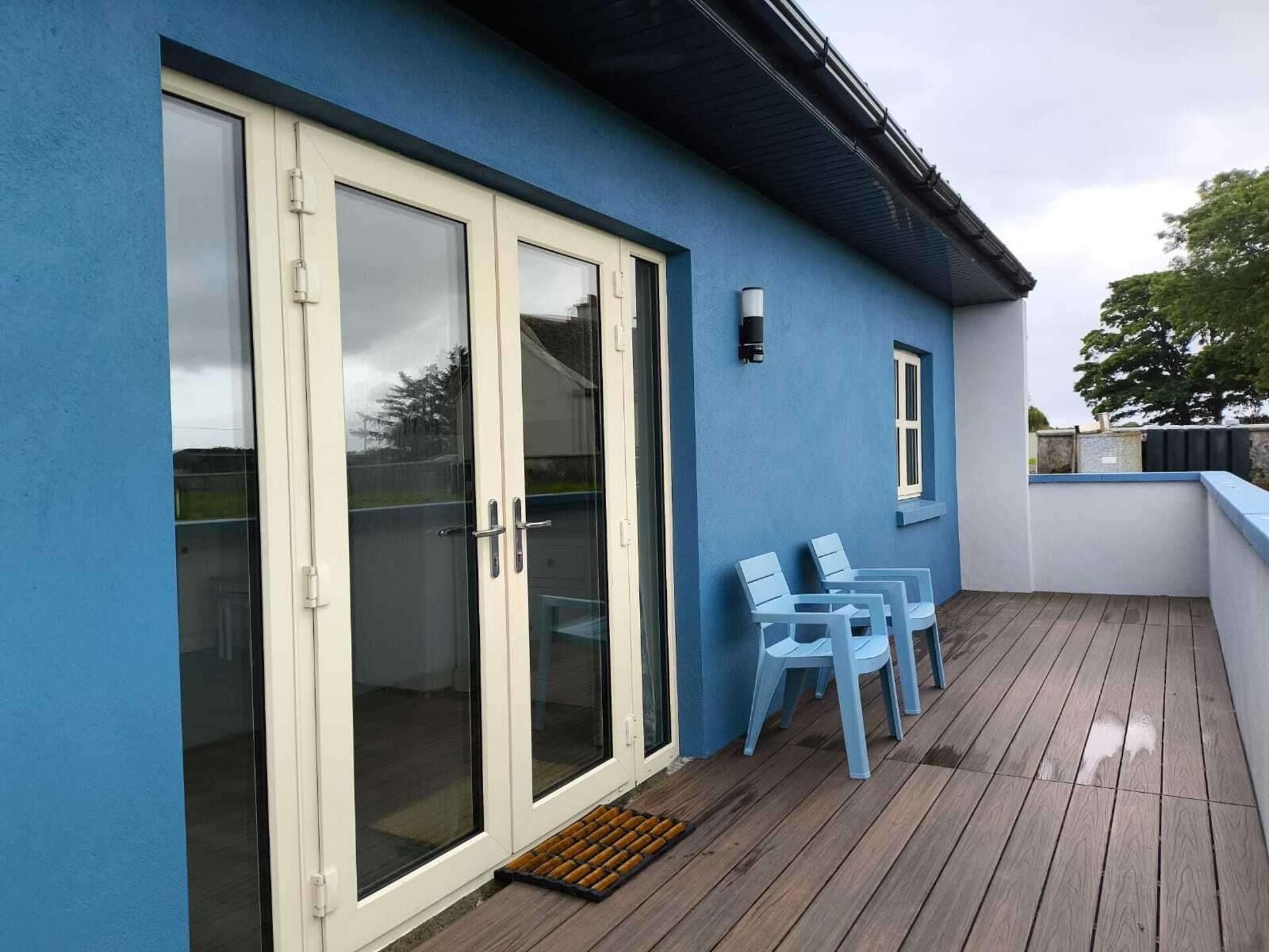 Un bâtiment bleu avec une porte coulissante en verre, une petite terrasse avec des chaises et une fenêtre.