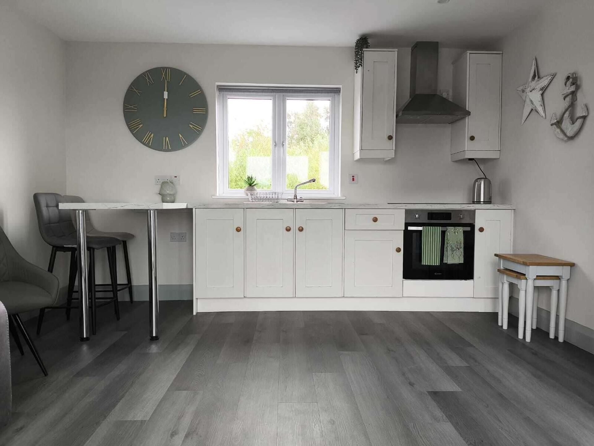 A modern kitchen with white cabinetry, a large wall clock, a window with a view, and a small dining area.