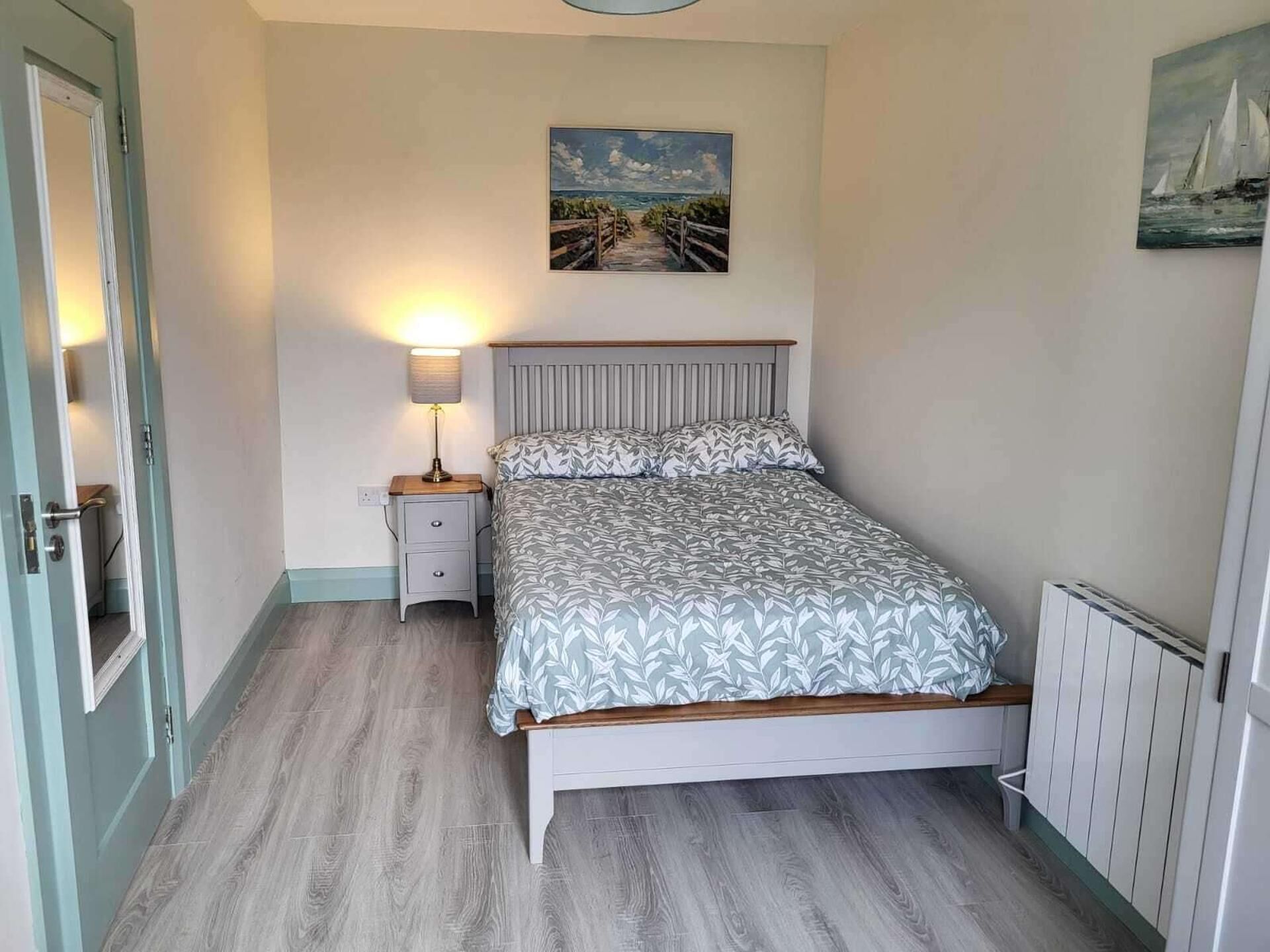 Un dormitorio con una cama, una mesita de noche con lámpara, un cuadro en la pared y una puerta.